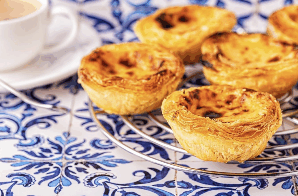 Pastel des Natas are on a blue tiled table next to a cup of coffee in Portugal