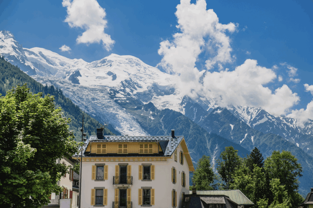 A yellow and white building stands before the snow covered French Alps on a sunny day