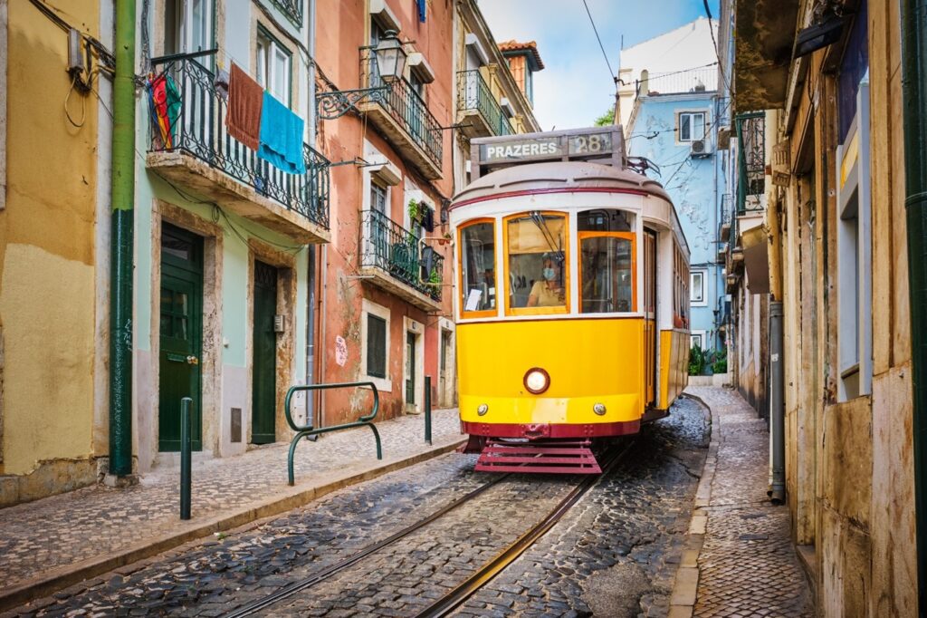 A classic yellow Lisbon tram traveling through a narrow cobblestone street, flanked by colorful buildings with balconies and hanging laundry in a historic neighborhood.