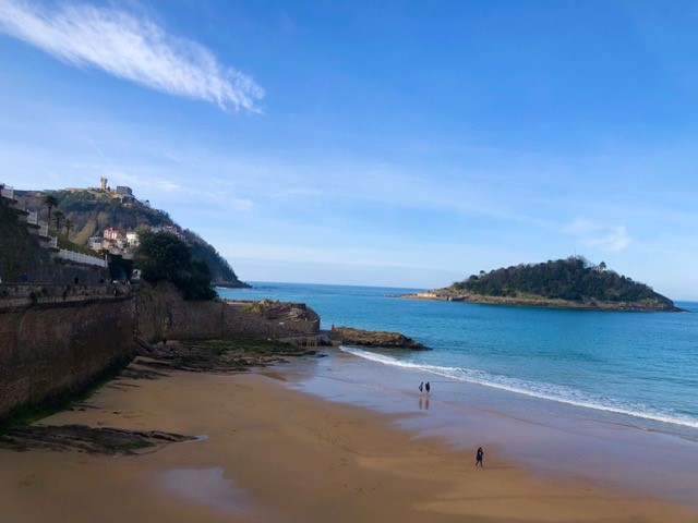 A quiet sandy beach in Spain’s Basque Country, with gentle waves, a few people walking along the shoreline, and green hills and coastal buildings framing the calm blue sea.