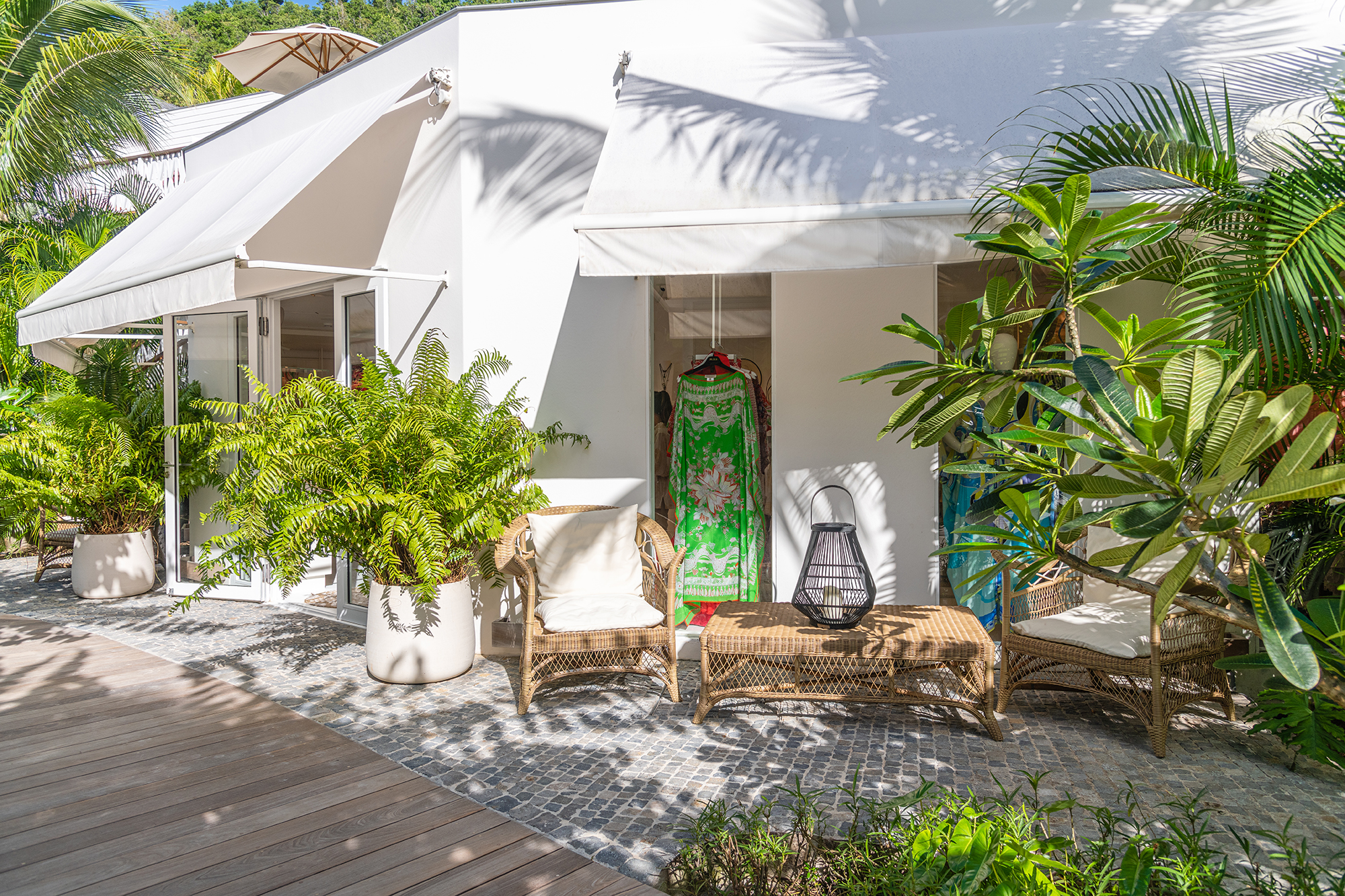 Chic tropical boutique exterior with white canvas awnings, lush green plants, woven seating, and a colorful dress displayed at the entrance, creating a serene, upscale island shopping atmosphere.