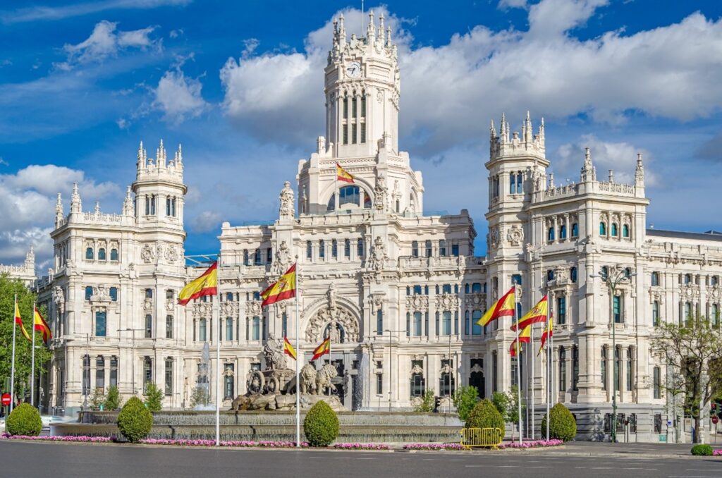 The Palacio de Cibeles in Madrid, a grand white civic building with ornate towers and Spanish flags, set along a wide plaza beneath a bright blue sky.