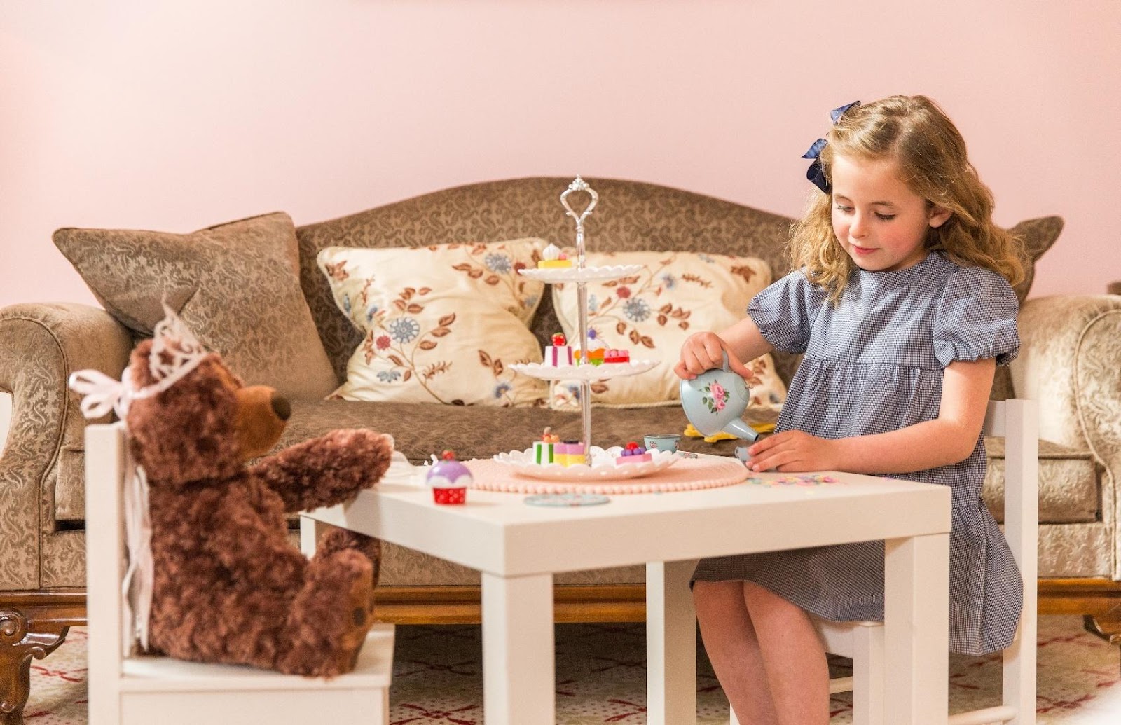 Young girl in blue gingham dress having a pretend tea party with teddy bear, pouring from floral teapot at small white table with tiered cake stand.