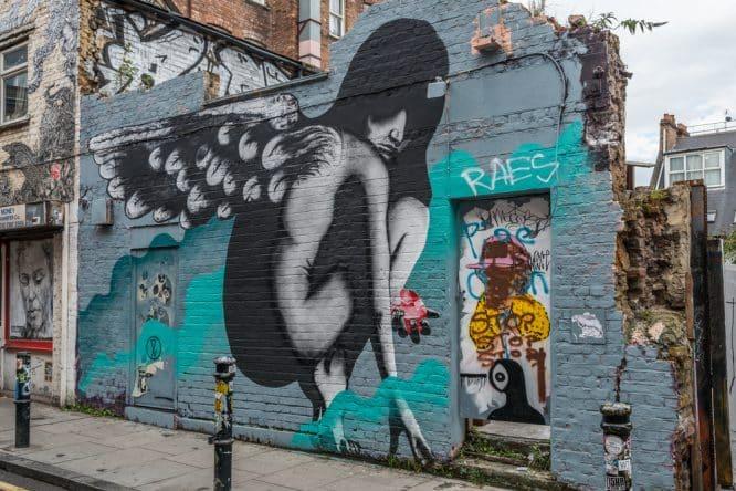 Street art mural of an angel with wings in black and white on turquoise blue brick wall in London with graffiti tags.