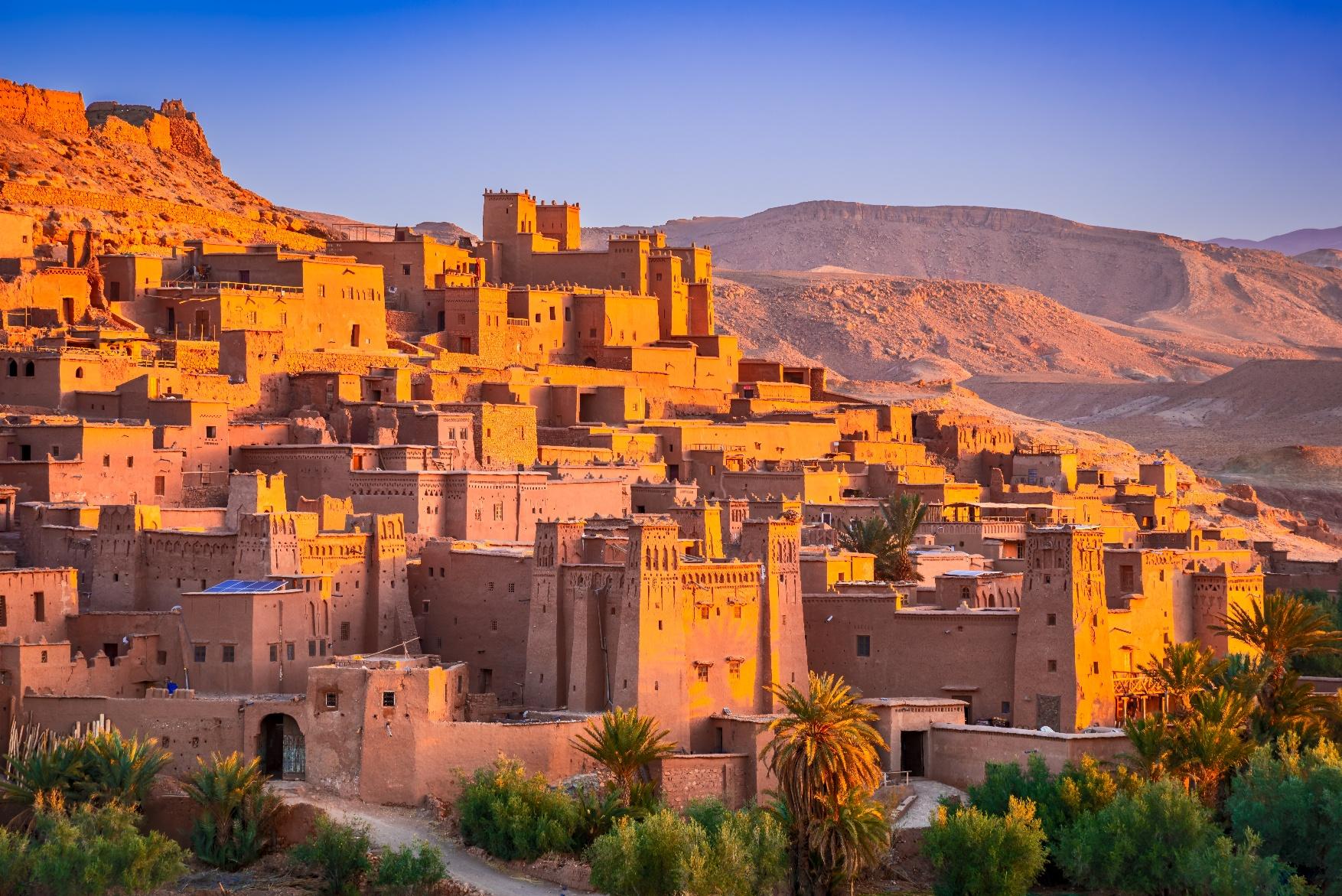 Luxury travel view of Ait Benhaddou in Morocco, a historic clay village glowing in warm desert light against the Atlas Mountains.